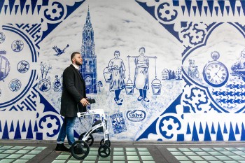 Man walking with a Rollz Motion Delft Blue rollator in front of a wall with drawings in Delft